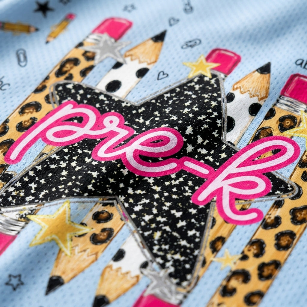 Close-up of a light blue fabric surface decorated with a star and pencil graphic. The central star is black with white stars and the word 'pre-k' in pink script, surrounded by illustrated pencils in leopard print, white with black spots, and pink erasers.