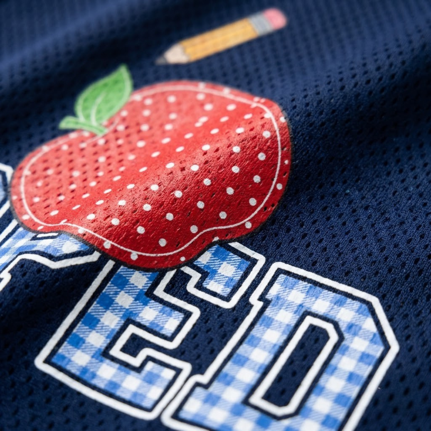 Macro close-up shot of the graphic print on mesh fabric, focusing on the high-definition blue gingham check pattern and red stitched apple texture.