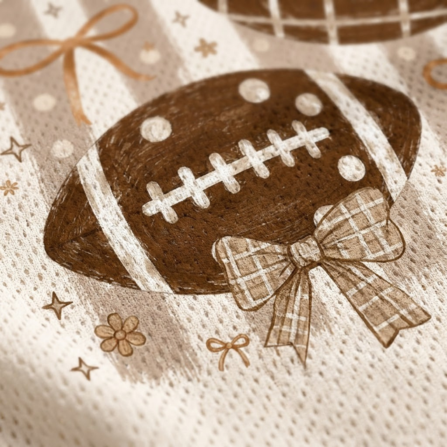 A close-up of a textured cream fabric featuring a brown football graphic with white laces, a brown plaid bow, and small gold stars and bows.