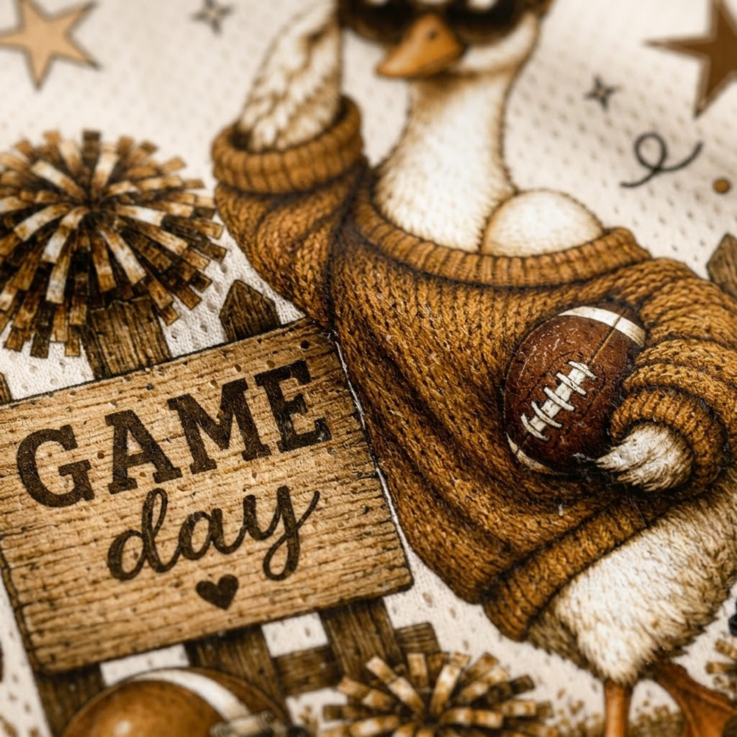 Close-up of a printed fabric or textile design featuring a duck in a brown sweater holding a football, with a wooden-style sign reading “GAME day” and autumn sports motifs.