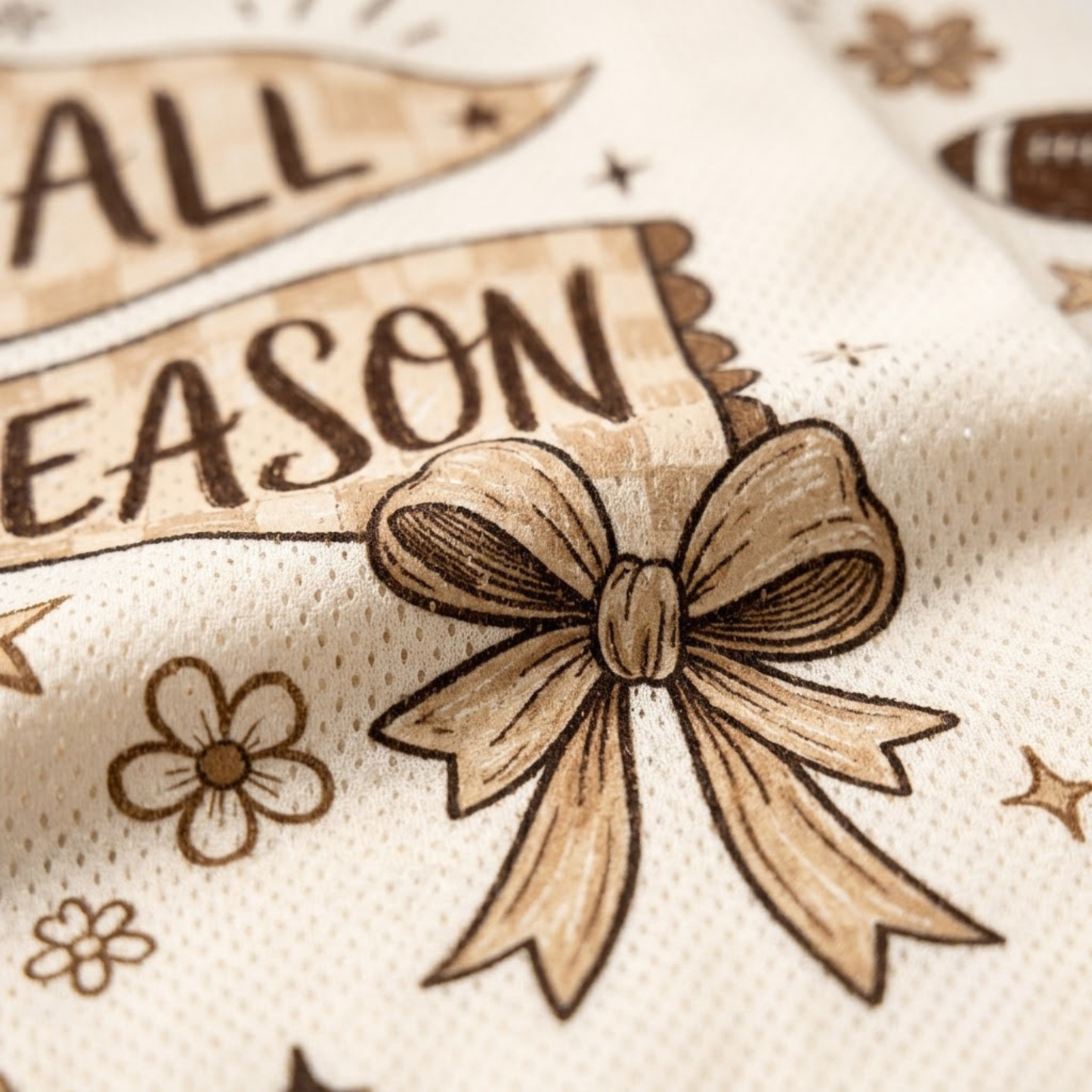 Close-up of a cream-colored fabric product featuring a brown printed design with the words 'ALL SEASON', stars, flowers, and a large bow motif.