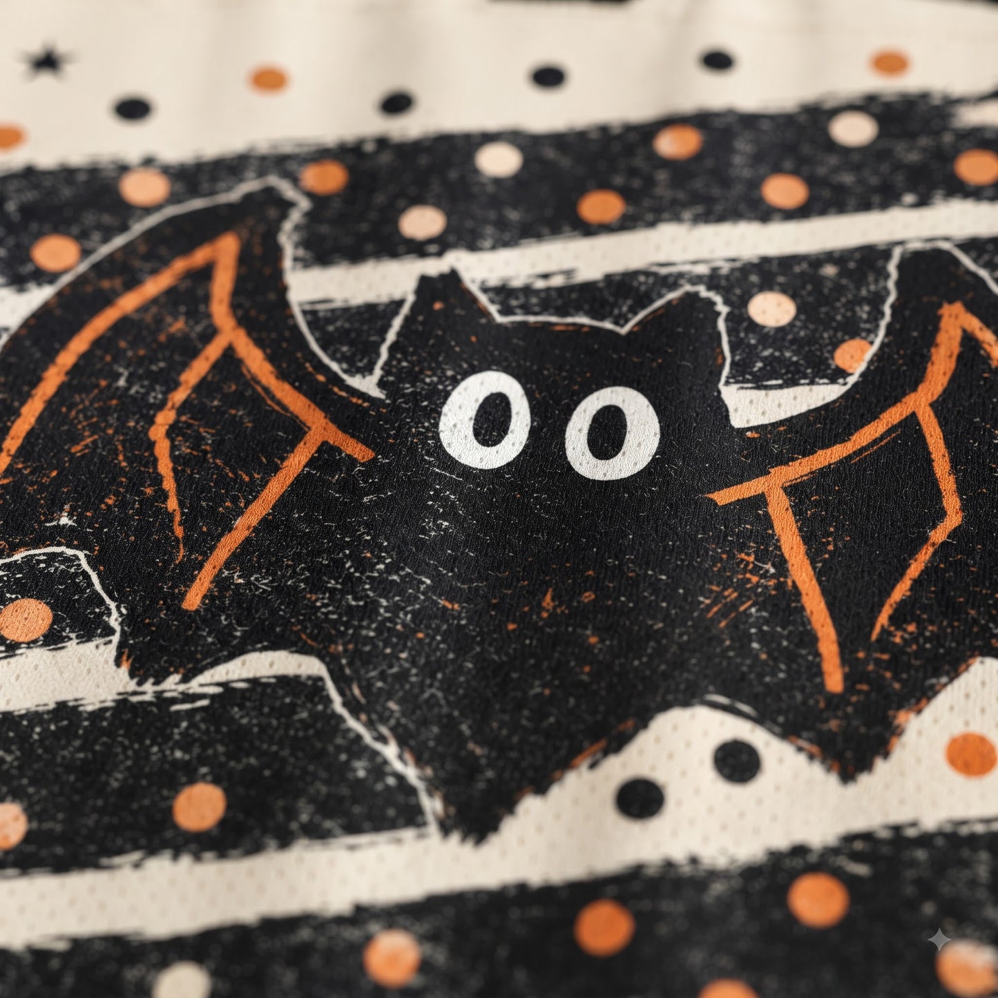 Close-up of a Halloween-themed fabric print featuring a black bat with orange wings and white eyes, surrounded by black and orange polka dots on a cream background.