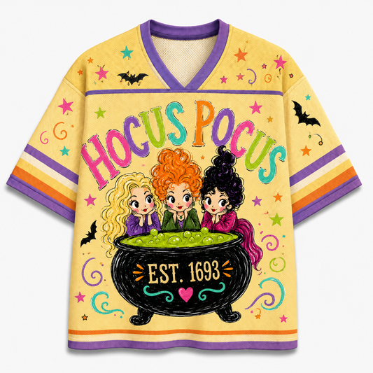 A short-sleeve V-neck t-shirt featuring a colorful 'Hocus Pocus' graphic with the three witch characters from The Three Brides sitting in a cauldron, set against a pale yellow background with purple and orange trim.