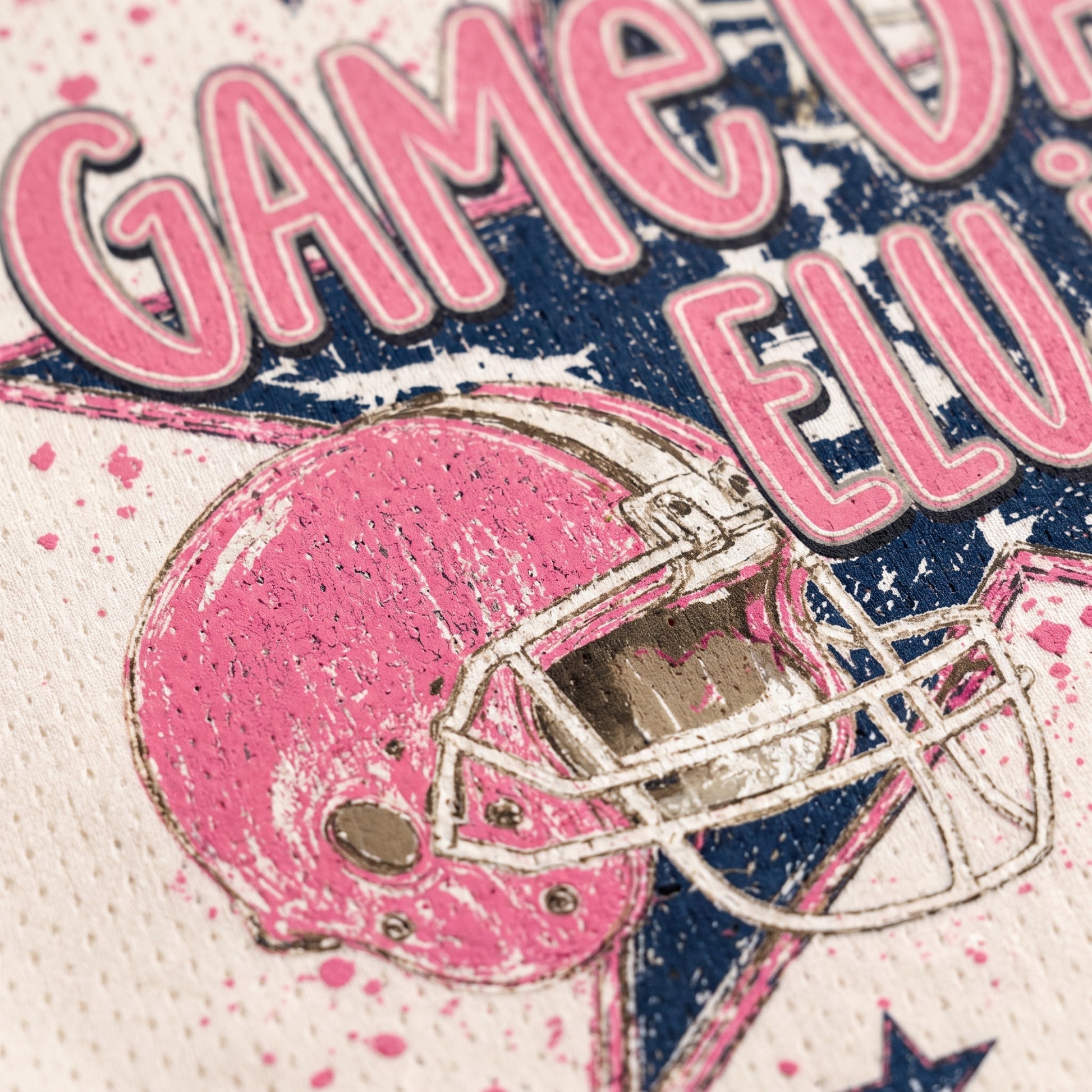 Close-up of a distressed graphic print featuring a pink football helmet with a white faceguard and large pink text reading 'GAME ON ELU' on a white background with pink and navy paint splatter details.