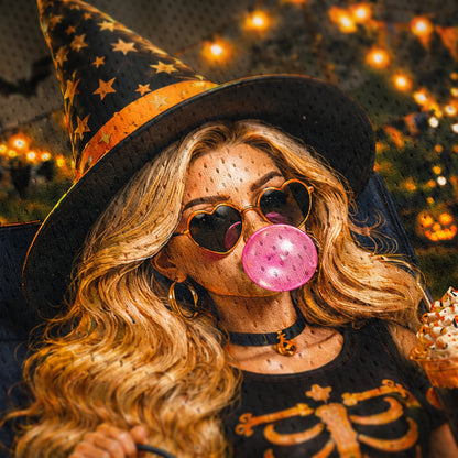 Person wearing a witch hat and skeleton shirt, blowing pink bubble gum with blurred lights in the background