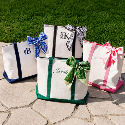 Four tote bags with monograms and colorful bows on a stone surface with grass in the background.