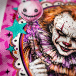 A close-up of a colorful printed design featuring a creepy clown face with white makeup, red blood-like streaks, orange hair, and a pink-and-white ruffled collar, holding a balloon with a stitched face. Teal stars and a bright pink patterned background surround the artwork.