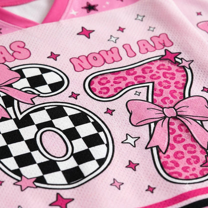 A close-up of a pink mesh garment, likely a sports jersey or top, featuring a graphic design with the text 'NOW I AM' and stylized numbers 0 and 1. The number 1 has a pink leopard print pattern with a pink bow, while the number 0 has a black and white checkerboard pattern.