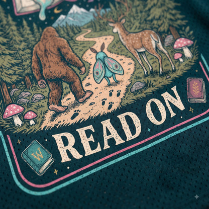 Close-up of a dark teal graphic t-shirt featuring a whimsical forest illustration with Bigfoot, a deer, a butterfly, mushrooms, books, and the text 'READ ON'.