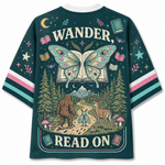 A dark teal graphic t-shirt shown from the back, featuring a whimsical illustration of a butterfly over an open book, a Bigfoot and deer in a forest, and the text 'WANDER, READ ON'. The sleeves have pink, white, and teal stripes.