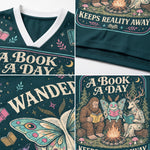 A dark teal graphic V-neck t-shirt shown in a four-panel collage, featuring the text 'A BOOK A DAY' and whimsical illustrations of a campfire, a butterfly, a big-footed creature, and a deer reading in a forest.