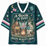 A dark teal V-neck graphic t-shirt featuring a whimsical forest illustration of a bear, a butterfly, and a deer reading by a campfire, with the text 'A BOOK A DAY KEEPS REALITY AWAY'. The sleeves have pink, white, and teal stripes, and the shirt is decorated with stars, moons, and mushrooms.