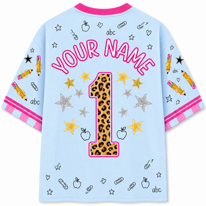 A light blue short-sleeve t-shirt with a customizable first birthday design, featuring a leopard-print number 1 outlined in pink, pink 'YOUR NAME' text, and school-themed doodles on a white background.