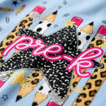 Close-up of a light blue fabric surface decorated with a star and pencil graphic. The central star is black with white stars and the word 'pre-k' in pink script, surrounded by illustrated pencils in leopard print, white with black spots, and pink erasers.