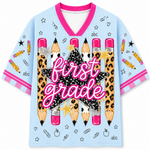 A light blue short-sleeve V-neck t-shirt with a pink collar and pink striped sleeve cuffs. The front features a large graphic of leopard-print pencils, a black glittery star, a gold star, and the words 'first grade' in pink script.
