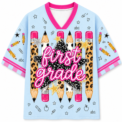 A light blue short-sleeve V-neck t-shirt with a pink collar and pink striped sleeve cuffs. The front features a large graphic of leopard-print pencils, a black glittery star, a gold star, and the words 'first grade' in pink script.