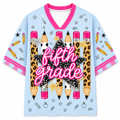 A light blue short-sleeve V-neck t-shirt with a 'fifth grade' school-themed graphic. The design features a large central black star with leopard print and pink glittery text, surrounded by illustrated pencils, stars, paperclips, and small 'abc' labels.
