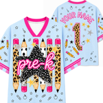 Front and back views of a personalized kids' t-shirt in light blue. The shirt features pink trim, a large central leopard-print star with the text 'pre-k' in pink, and illustrations of pencils, stars, apples, and paperclips.
