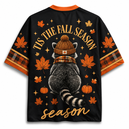 A black short-sleeve t-shirt with an all-over autumn-themed graphic print. The design features a raccoon wearing a brown knit beanie and plaid scarf, surrounded by orange maple leaves, pumpkins, stars, and the text 'TIS THE FALL SEASON season'.