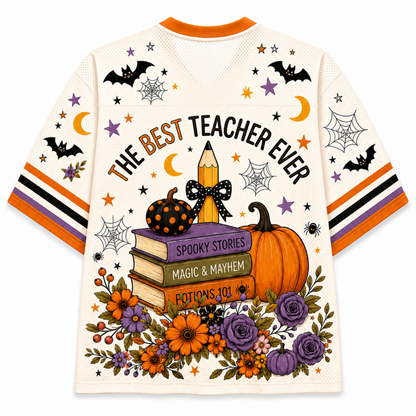A cream-colored mesh short-sleeve t-shirt with an orange collar, decorated with a colorful Halloween theme. The back of the shirt features the text 'THE BEST TEACHER EVER' arched over a stack of books, a polka-dot pumpkin, a pencil, and a large floral arrangement at the bottom.
