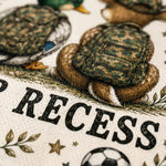 Close-up of a printed textile or fabric featuring illustrated ducks wearing camouflage backpacks, large black text reading 'RECESS,' and small decorative motifs like leaves, stars, and a soccer ball on an off-white background.