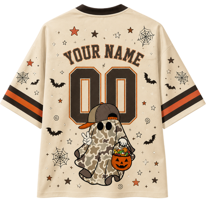 A beige short-sleeve t-shirt with a black collar, featuring a Halloween-themed graphic on the back. The design includes 'YOUR NAME' and a customizable number '00' in dark brown and orange, a camouflage-patterned ghost holding a pumpkin bucket, and various spiderwebs, bats, and stars.