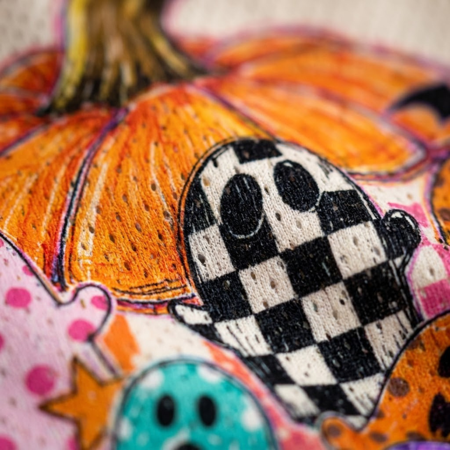 Close-up of a colorful embroidered design featuring orange pumpkins and black-and-white checkered ghosts on a textured fabric background.