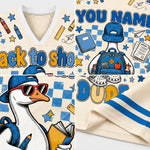 A close-up collage of a cream-colored athletic jersey or sweatshirt fabric with blue and yellow printed graphics. The design includes a V-neck, mesh texture, blue stripes, a cartoon duck wearing a blue cap and sunglasses with a backpack, stars, pencils, books, and text such as 'YOU NAME' and 'DUDS'.