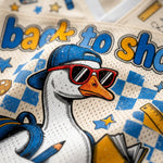 Close-up of a graphic t-shirt featuring a cartoon duck wearing a blue cap and red sunglasses, with 'back to school' text and school-themed doodles on a cream mesh fabric.