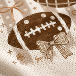 A close-up of a textured cream fabric featuring a brown football graphic with white laces, a brown plaid bow, and small gold stars and bows.