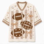 A short-sleeve V-neck athletic-style top in cream with brown and tan football graphics, striped vertical panels, and small bow and star motifs, shown on a plain light background.