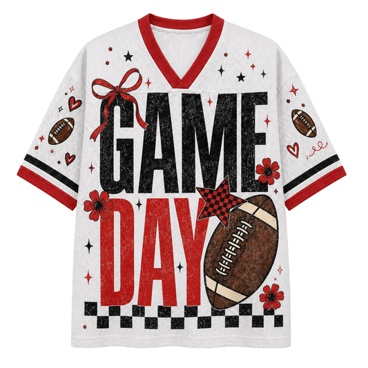 A white short-sleeve V-neck jersey-style shirt with red trim and a distressed 'GAME DAY' graphic, featuring footballs, stars, hearts, flowers, and a checkered stripe at the bottom.