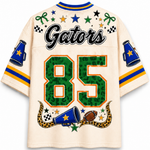 A cream-colored short-sleeve jersey with 'Gators' and the number '85' printed on the back. The jersey features green leopard-print lettering for the number 85, blue and yellow stripes on the sleeves, and various sports-themed graphics including stars, checkered flags, a football, and a snake.