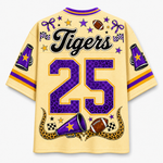 A short-sleeve t-shirt shown from the back, featuring a light yellow base with purple and gold graphics. The back is decorated with the word 'Tigers' in black script, the number '25' in purple leopard print with gold borders, and football-themed illustrations including a football, megaphones, stars, and checkered flags.