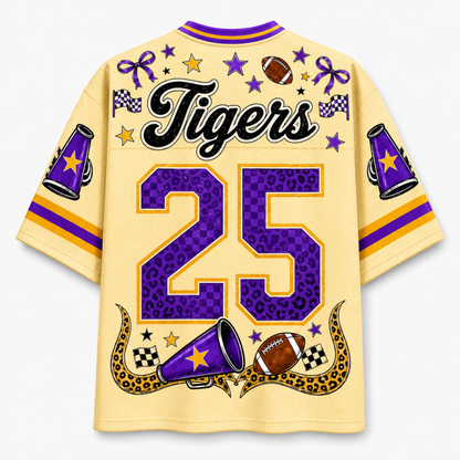 A short-sleeve t-shirt shown from the back, featuring a light yellow base with purple and gold graphics. The back is decorated with the word 'Tigers' in black script, the number '25' in purple leopard print with gold borders, and football-themed illustrations including a football, megaphones, stars, and checkered flags.