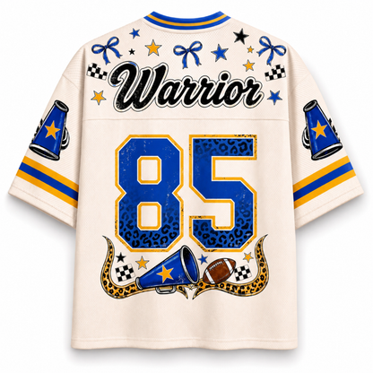 A cream-colored short-sleeve jersey shown from the back, featuring 'Warrior' in black script across the upper back, the number '85' in blue with leopard-print detailing, and blue, yellow, and black graphic accents including stars, bows, checkered flags, and a football.