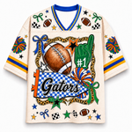 A short-sleeve V-neck graphic t-shirt in cream with blue and yellow accents, featuring a football-themed design with the word 'Gators' and various sports motifs like a football, foam finger, and stars.
