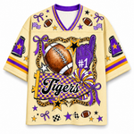 A short-sleeve athletic jersey with a purple and gold checkered V-neck collar, featuring a large football-themed graphic on the front with the word 'Tigers', a purple foam finger, stars, and decorative elements on a cream background.