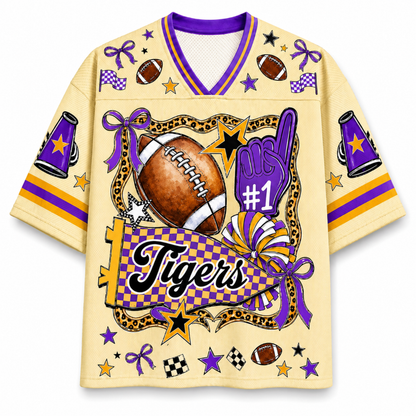 A short-sleeve athletic jersey with a purple and gold checkered V-neck collar, featuring a large football-themed graphic on the front with the word 'Tigers', a purple foam finger, stars, and decorative elements on a cream background.