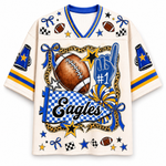 An off-white short-sleeve V-neck jersey with blue and yellow athletic stripes on the sleeves and collar, featuring a large football-themed graphic print with the word 'Eagles', a football, stars, bows, and a blue 'No. 1' finger graphic.