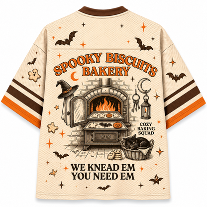 A beige short-sleeve graphic T-shirt shown from the back, featuring Halloween-themed illustrations and text including 'Spooky Biscuits Bakery,' a bakery oven scene, bats, stars, and the phrase 'We knead em you need em.' The shirt has brown and orange sleeve stripes and a brown collar.