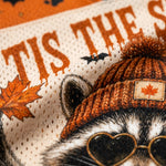 Close-up of a knit beanie with a maple leaf patch, shown on a raccoon illustration with heart-shaped sunglasses and autumn-themed graphics.