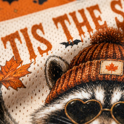 Close-up of a knit beanie with a maple leaf patch, shown on a raccoon illustration with heart-shaped sunglasses and autumn-themed graphics.
