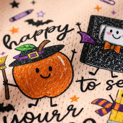 Close-up of a peach-colored fabric printed with a Halloween-themed design, featuring a smiling orange pumpkin wearing a black witch hat, a black book character with a purple bow tie, stars, bats, and partial black script text.