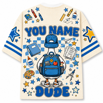 A cream-colored t-shirt with a large blue and yellow graphic print on the back. The design features a cartoon duck wearing a blue baseball cap and backpack, surrounded by school-themed doodles like stars, pencils, and a paint palette.