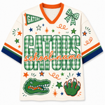 A Florida Gators themed short-sleeve jersey with a cream base, green and white gingham pattern, orange trim, and collegiate graphics including an alligator head, paw print, stars, and the word 'GATORS' across the front.