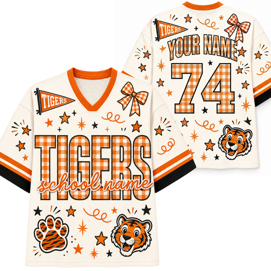 A custom tiger-themed football jersey shown from both front and back. The jersey has an off-white base with orange and black accents, featuring 'TIGERS' in large orange-and-white plaid letters, a tiger mascot graphic, a paw print, stars, and decorative bows.