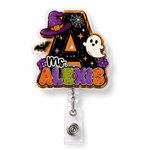 A Halloween-themed retractable badge reel featuring a die-cut letter 'A' with 'Ms. ALEXIS' text, decorated with a purple witch hat, white ghost, bats, and purple flowers on a black background.