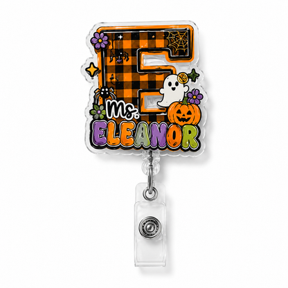 A Halloween-themed retractable badge reel featuring a die-cut acrylic face with the text 'Ms. ELEANOR' in colorful letters, decorated with a large orange-and-black buffalo plaid letter 'E', a ghost, a pumpkin, spiderwebs, and small purple flowers.