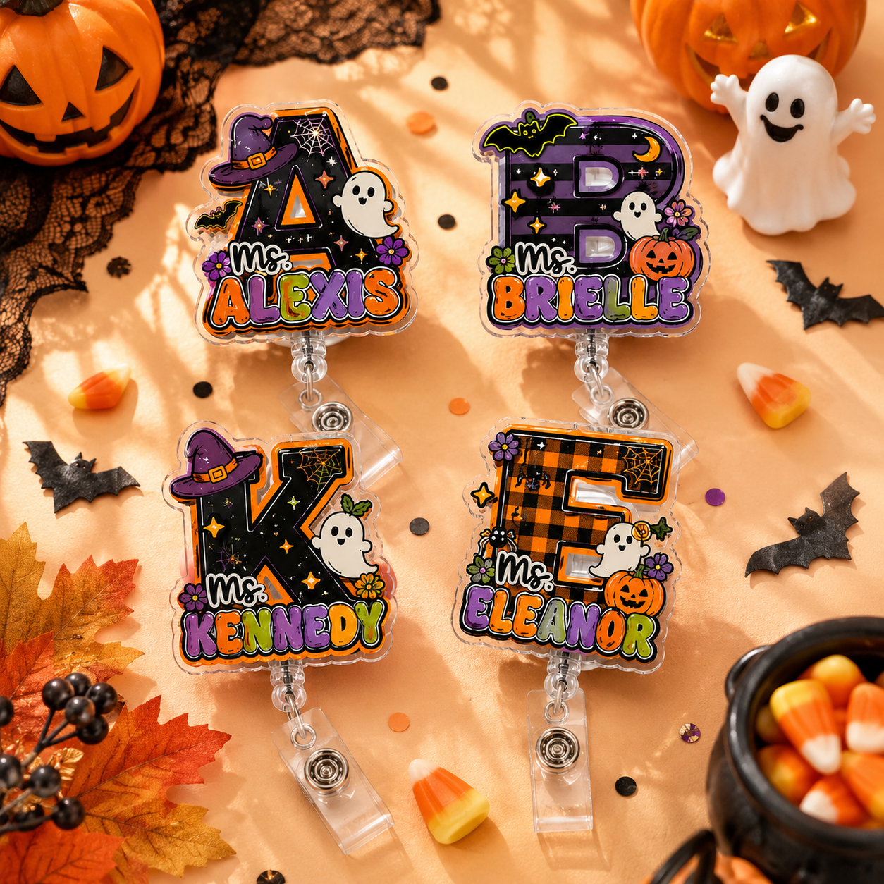 Four Halloween-themed acrylic name badges with clear plastic clip-on backs, decorated with letters A, B, K, and F, ghosts, pumpkins, bats, and autumn leaves on an orange fabric background with seasonal props.